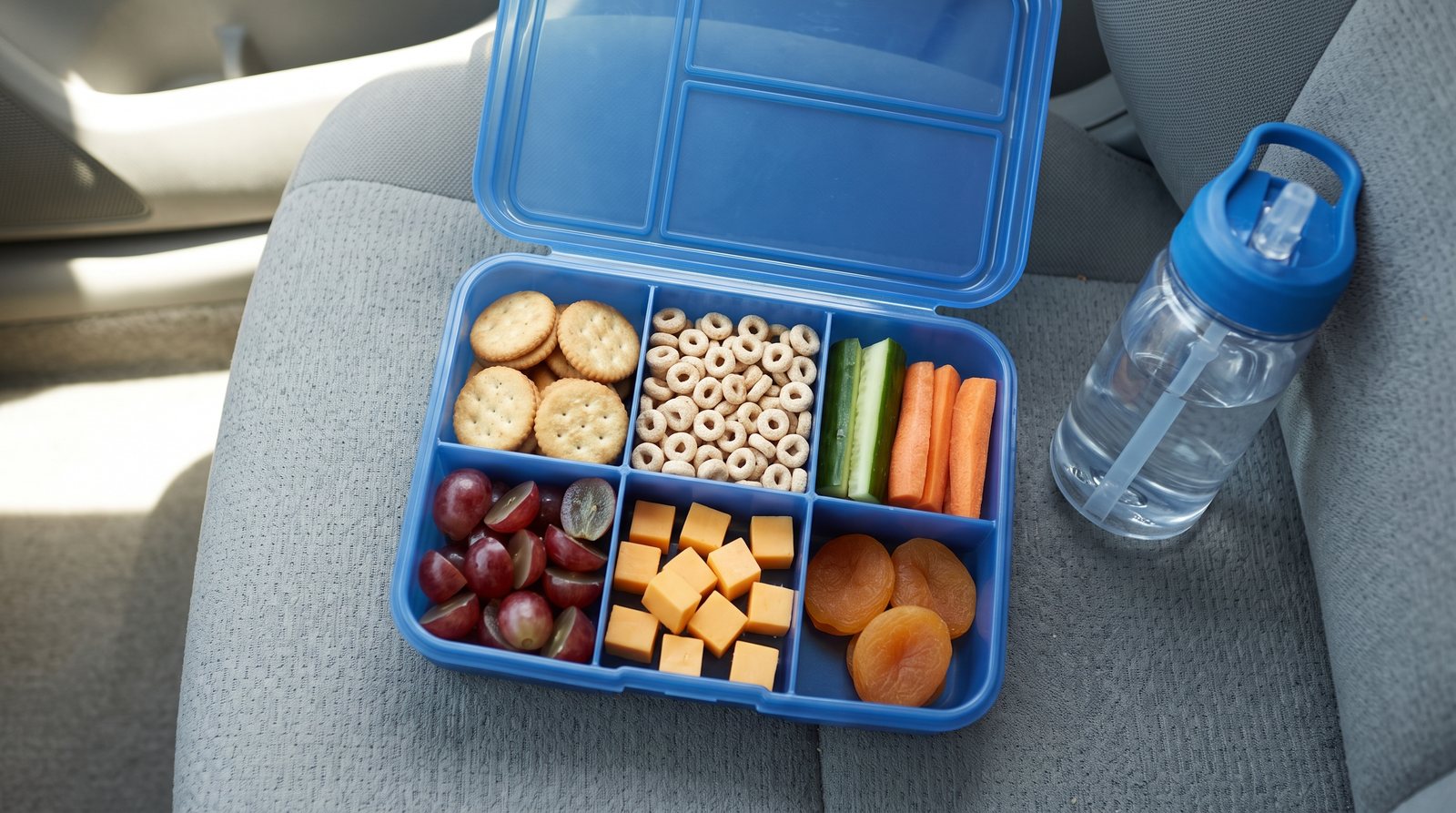 A blue divided snack box packed for a car trip with crackers, cereal, cucumber and carrot sticks, grapes, cheese cubes and dried apricots.